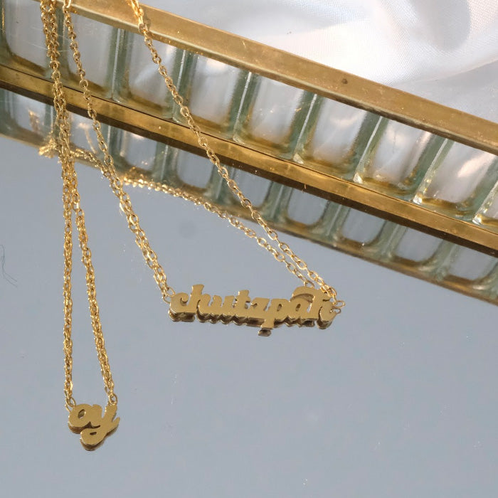 Close product shot of chutzpah gold necklace on mirror. There is also an Oy necklace next to it.