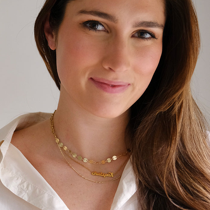 Woman with brown hair smiles at camera while wearing layered gold necklaces and white blouse.