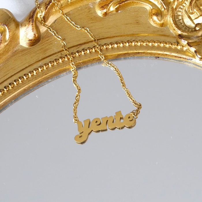 Product shot of gold yente nameplate necklace on gold ornate mirror.